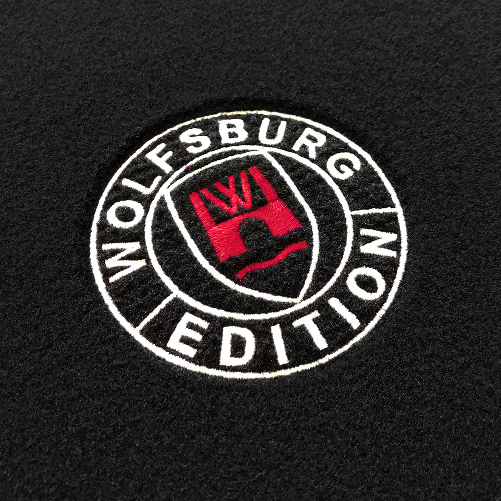 wob Stick Detail-103 rot Das gestickte Wolfsburg Editions Logo auf einem schwarzen Teppich und das Logo ist in rot.