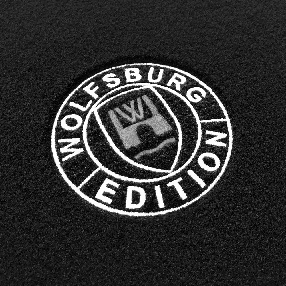 wob Stick Detail-502 grau Das gestickte Wolfsburg Editions Logo auf einem schwarzen Teppich und das Logo ist in grau.