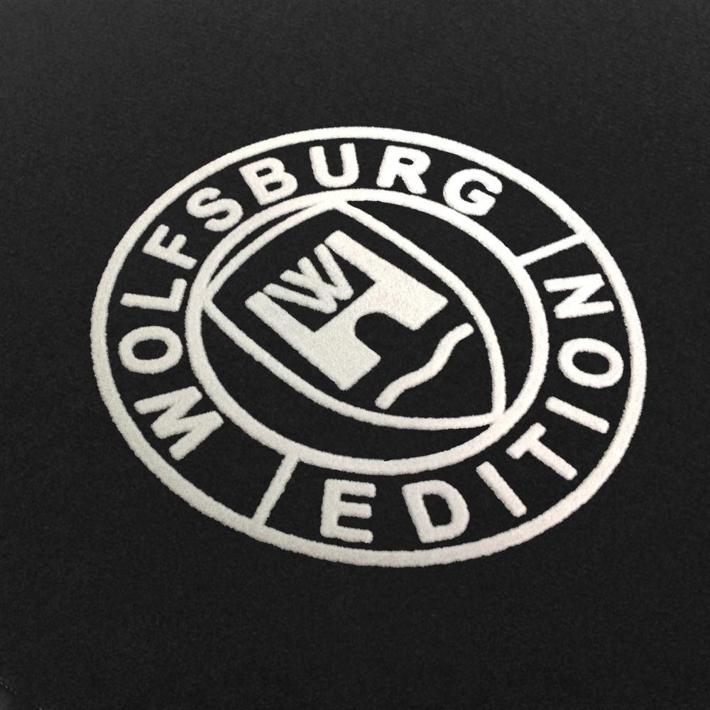 WOB-Detail-100 weiss Das Wolfsburg Editions Logo auf einem schwarzen Teppich und das Logo ist in weiss.