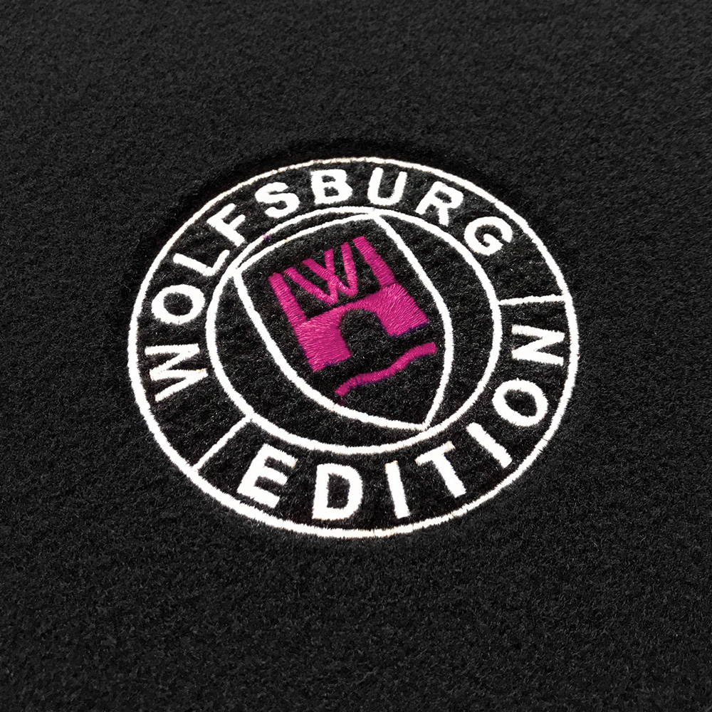 wob Stick Detail-404 pink Das gestickte Wolfsburg Editions Logo auf einem schwarzen Teppich und das Logo ist in pink.